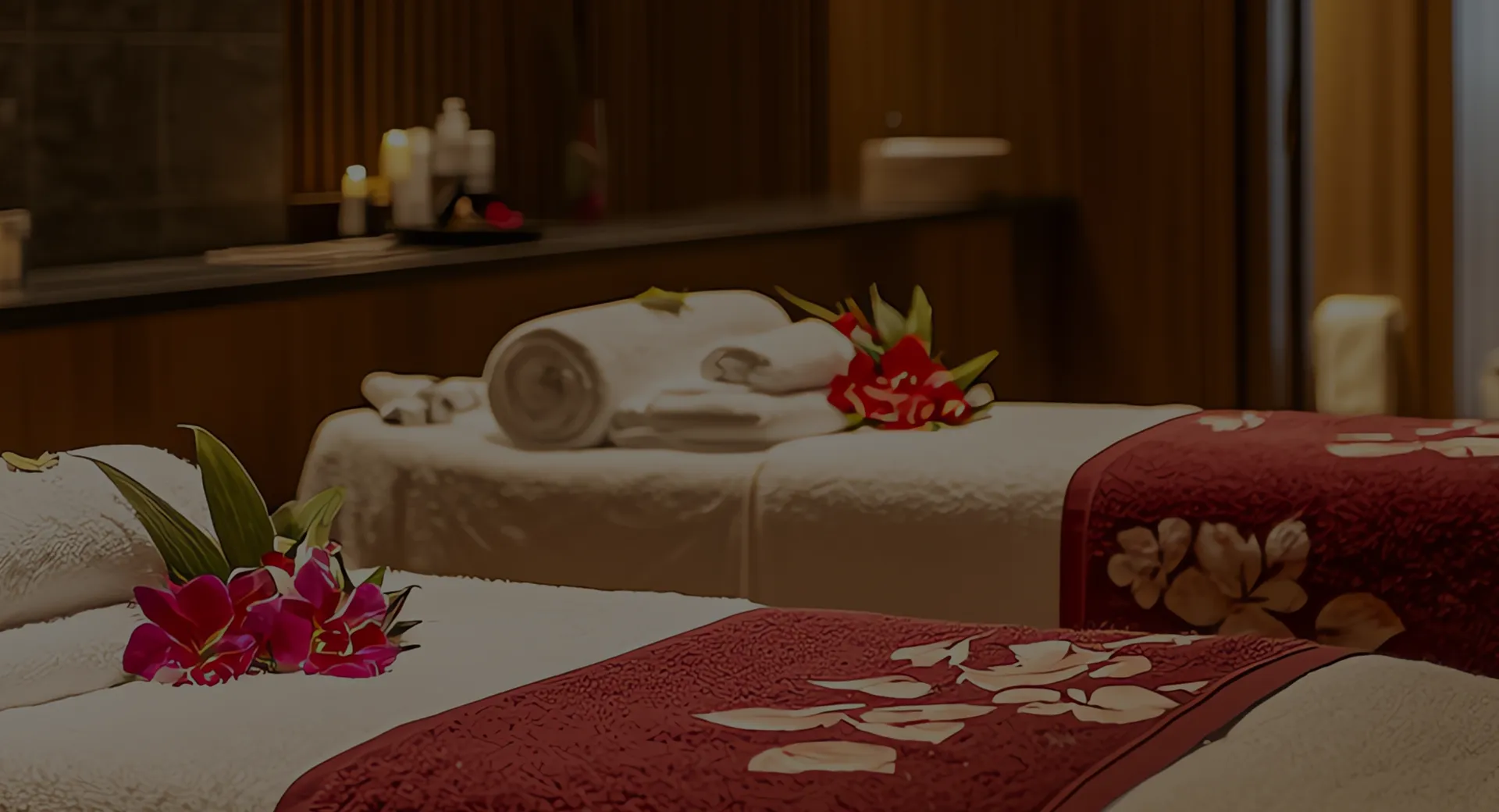 Two spa beds with red floral-patterned covers, white towels, and pink flowers in a warmly lit room.