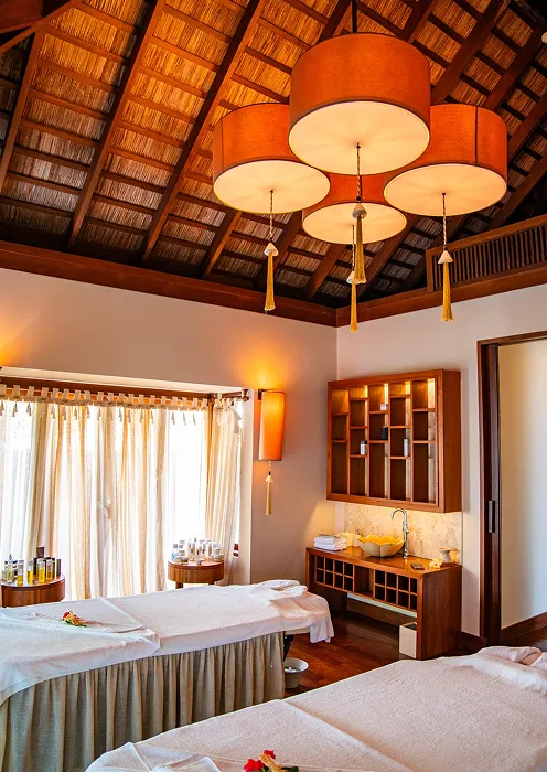 Spa room with two massage beds, a wooden ceiling, warm lighting, and shelves with towels and spa products.